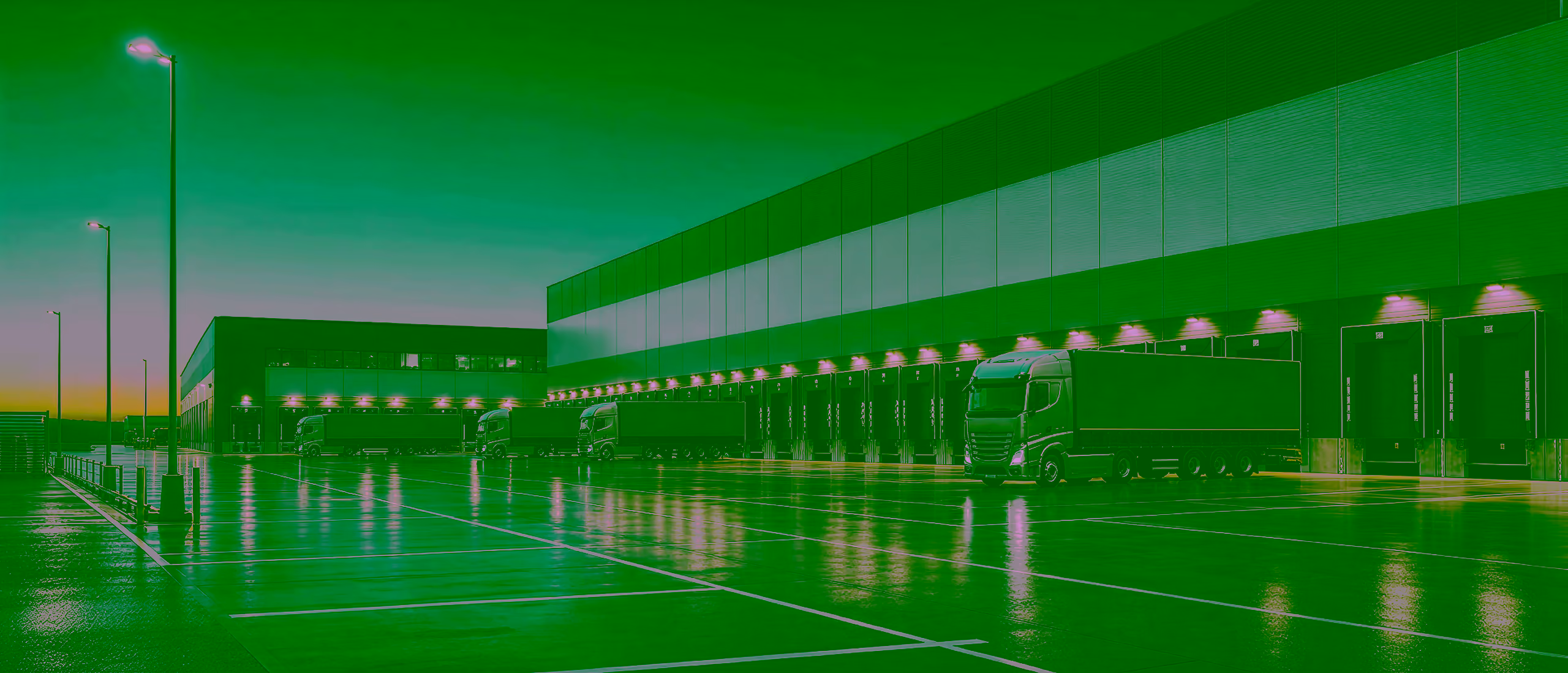 A distribution terminal at dusk: tractor units backed into a row of lit loading docks, their curtainsiders in line along a wet apron.